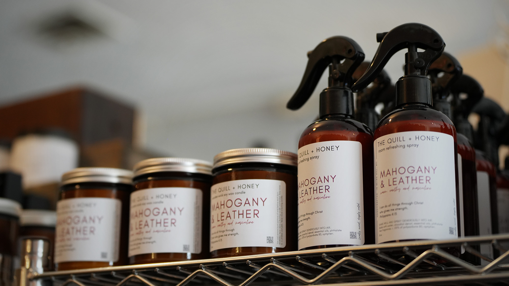 Jars and spray bottles labeled Mahogany & Leather by The Quill + Honey are displayed on a metal wire shelf, captured as part of our photography & videography services. The products feature brown and white labels with black caps or spray tops.