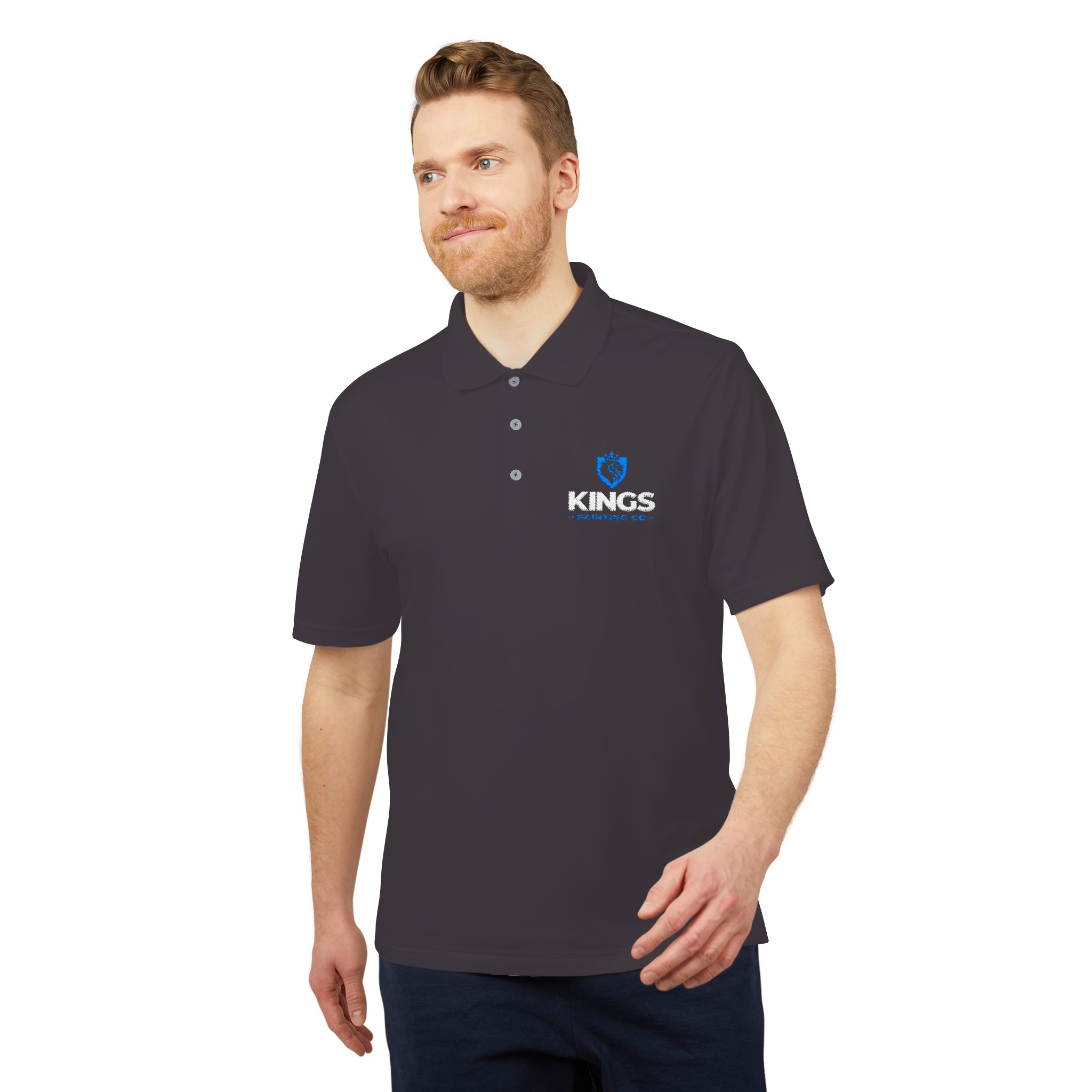 A man with light brown hair and a beard is standing and smiling slightly, wearing the Kings Painting Co. Performance Polo by Under Armour®, which features the KINGS logo on the left chest. His right hand rests relaxed by his side.